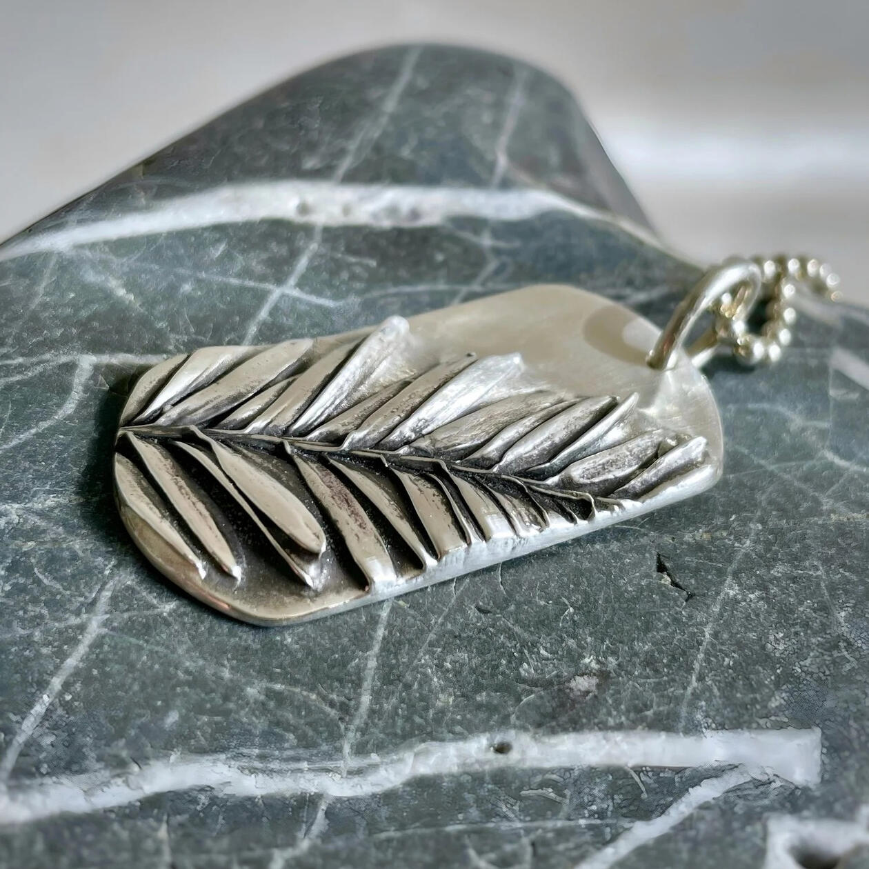 A sterling silver "dog tag" pendant rests on a striated stone. A silver redwood leaf seems to emerge in three dimensions from the face of the dog tag. Jessica made this!